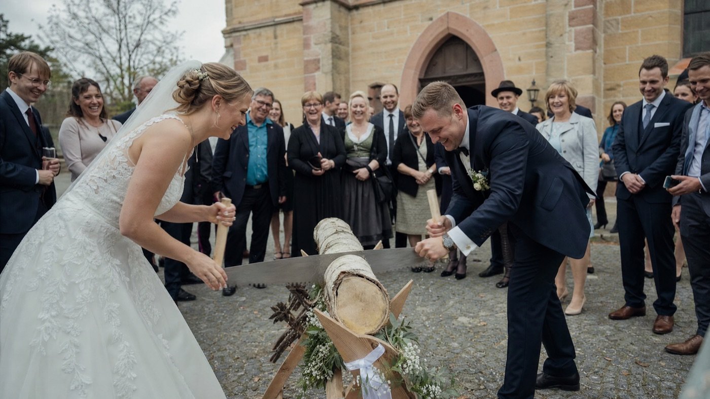 Brautpaar sägt gemeinsam einen Baumstamm vor der Kirche